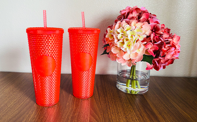 Two Red Starbucks Tumblers Next To a Vase with Flowers